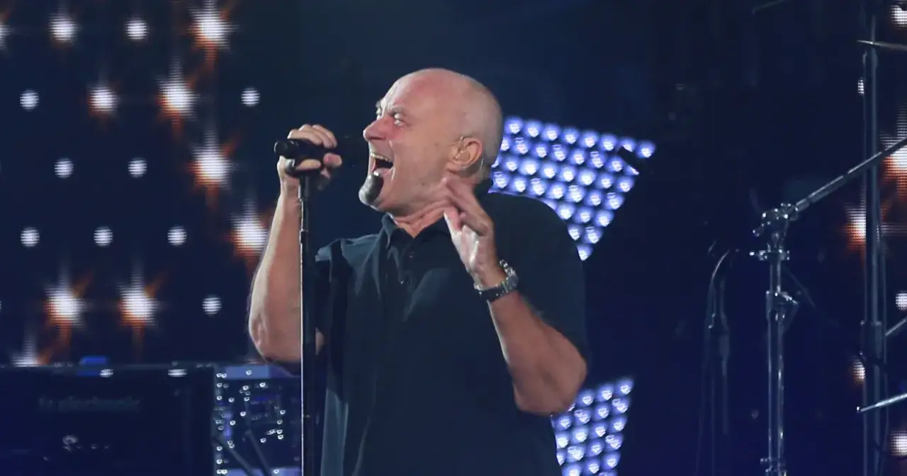 Phil Collins, mimo choroby, śpiewa na koncercie. Wokalista z mikrofonem na tle świateł.