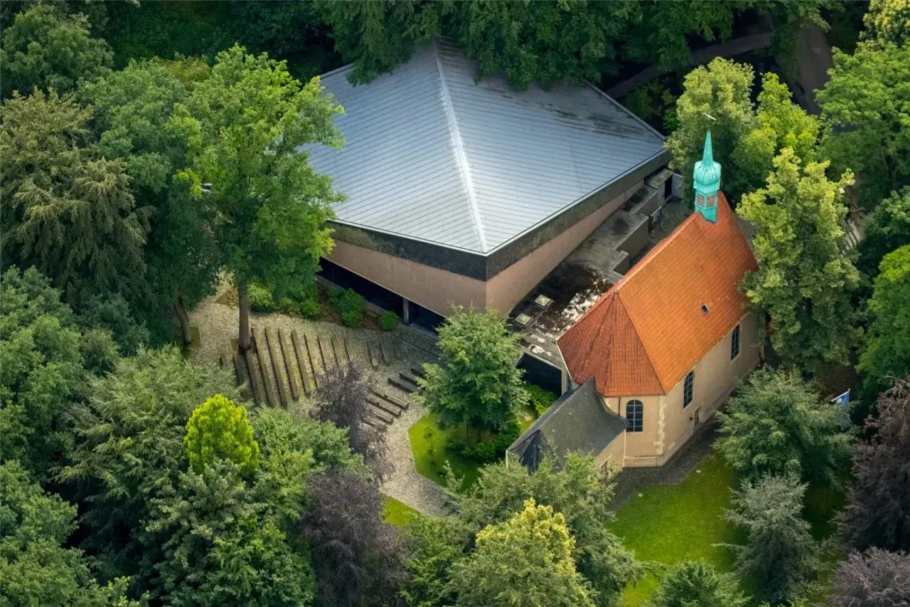 Luftaufnahme des Annabergs in Haltern, umgeben von üppigem Grün. Ein moderner Bau mit Pyramiden-Dach und eine traditionelle Kirche mit Ziegeldach sind zu sehen.