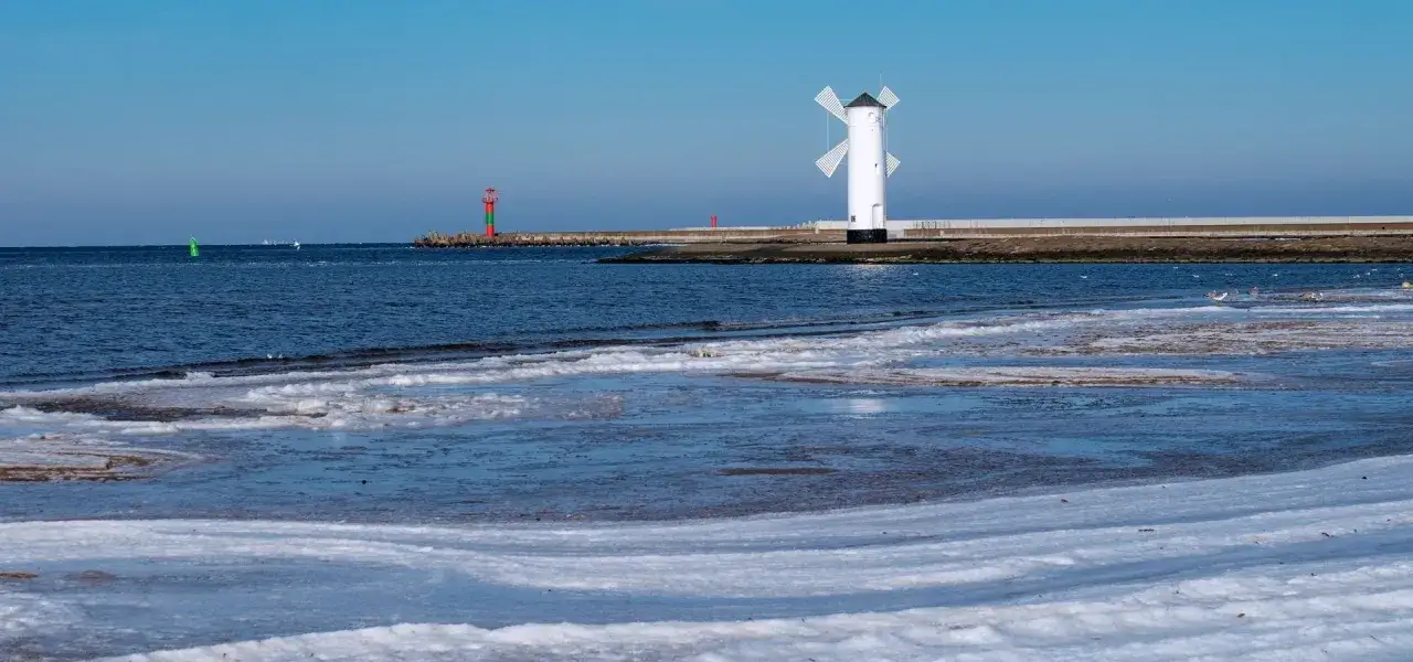 Zimowe świnoujście atrakcje: biały wiatrak-latarnia morska i czerwono-zielona latarnia na falochronie, zamarznięte fale na plaży.