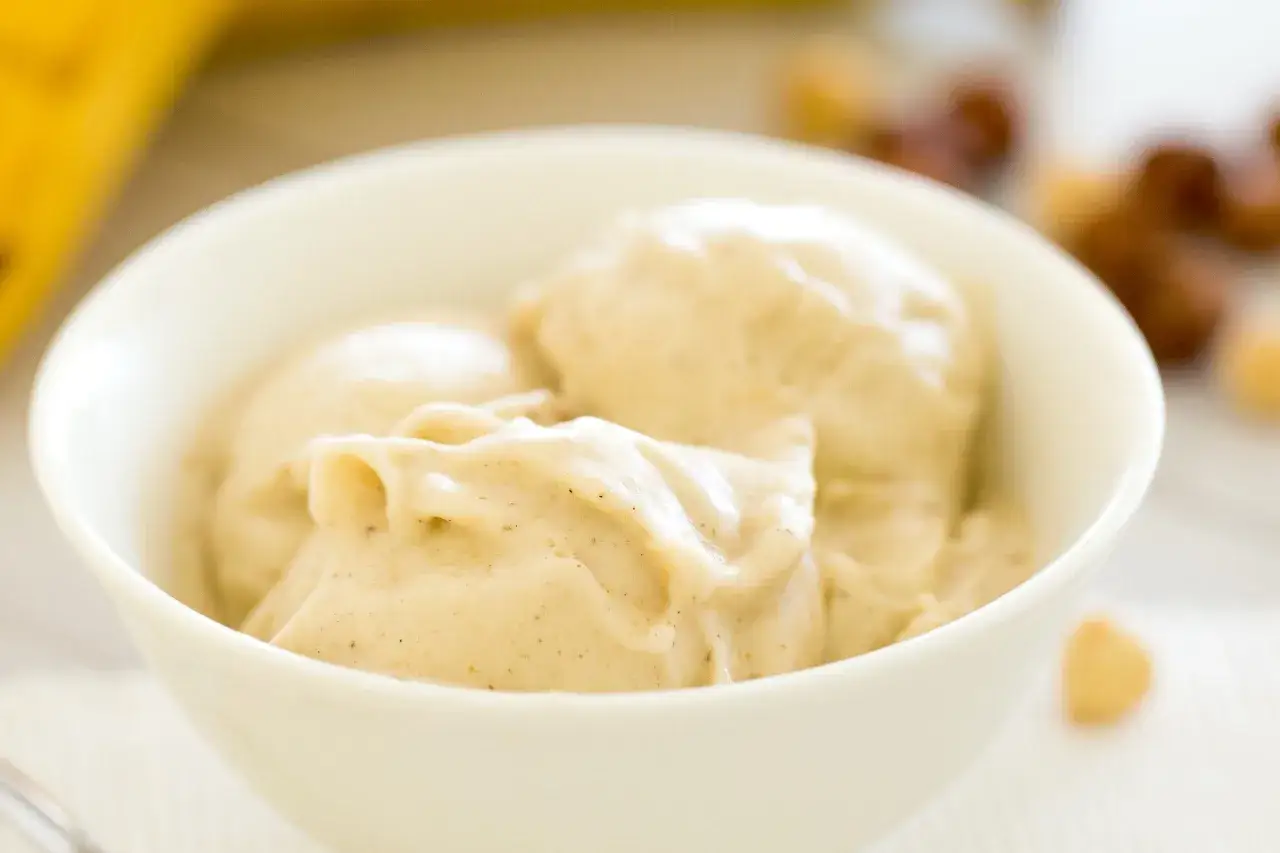Un cremoso helado de plátano en un cuenco blanco, con trocitos de plátano y frutos secos desenfocados al fondo.