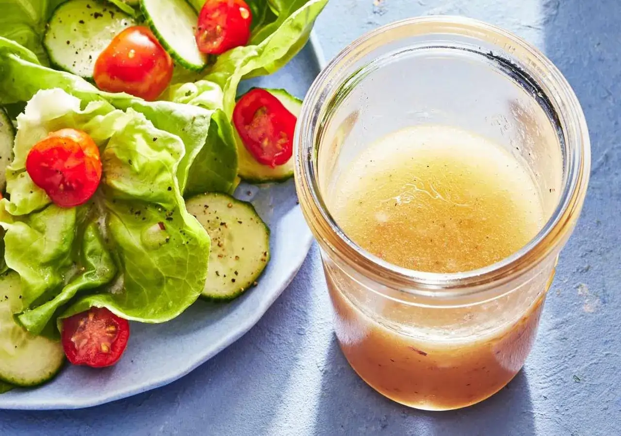 En burk med hemlagad salladsdressing bredvid en fr&auml;sch sallad med gurka och tomater. Ett perfekt salladsdressing recept f&ouml;r en god m&aring;ltid.