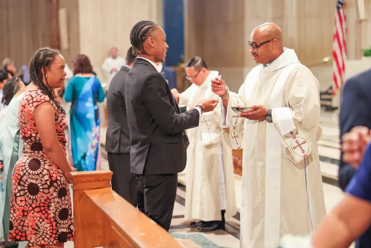 A deacon in the church, wearing a white stole, offers communion to a young man in a suit.