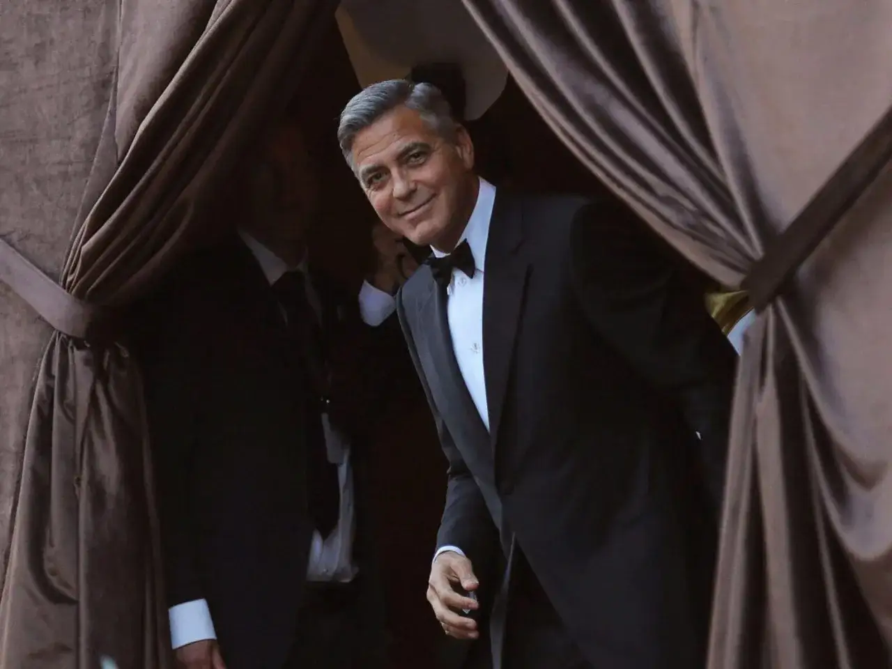 George Clooney, looking dapper in a classic tuxedo, emerges from behind a draped curtain.
