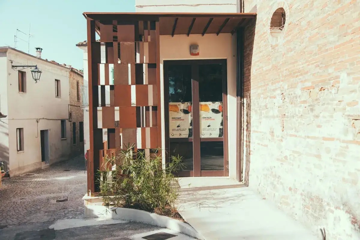 Ingresso moderno con pannelli metallici decorativi, un'esposizione artistica e un vicolo acciottolato che porta a San Ginesio.