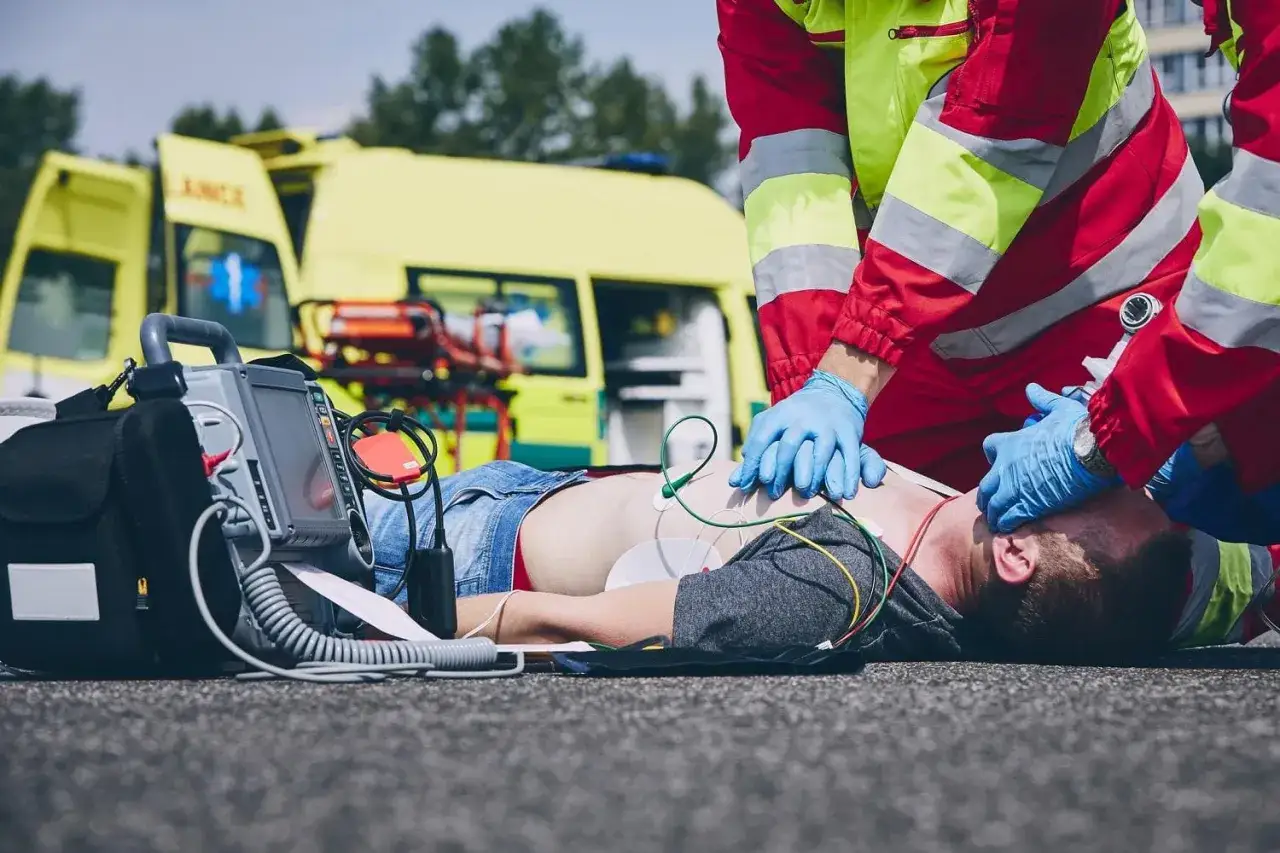 Ratownicy medyczni prowadzą resuscytację krążeniowo-oddechową. Obok leży defibrylator, a w tle żółta karetka. To przykład profesjonalnego abc ratownictwo.