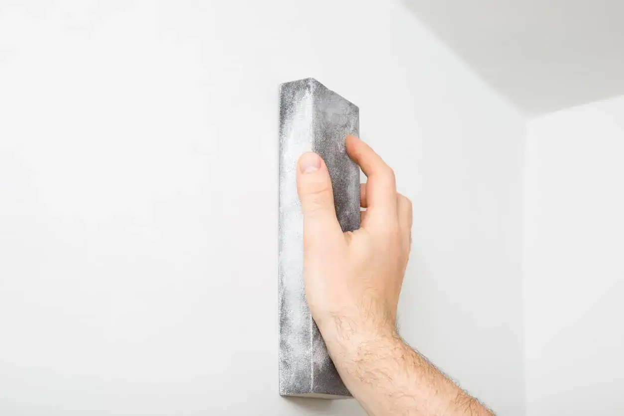 Mano lijando una pared blanca con un bloque de lijado. Se usa para alisar la masilla para techo escayola.