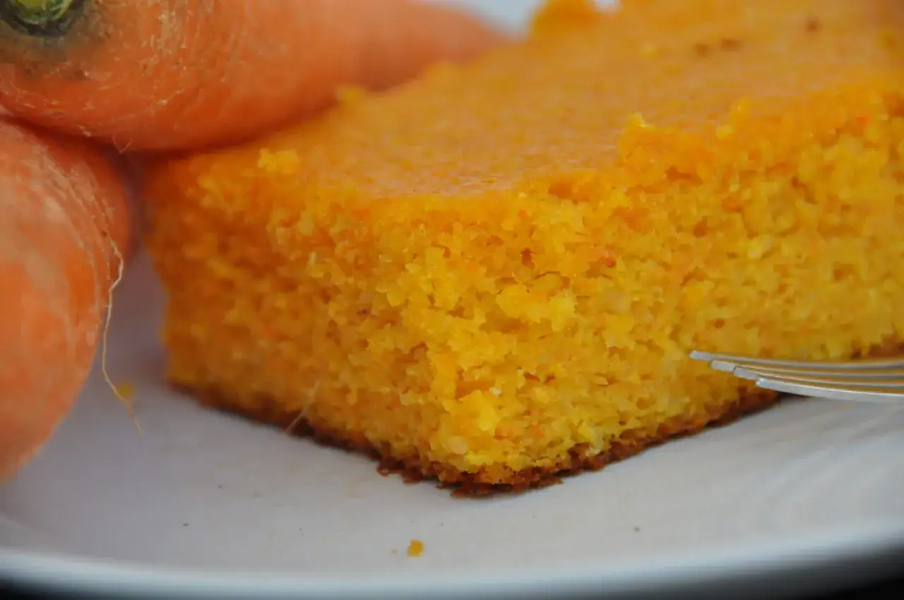 Una fetta di torta di carote fatta con il Bimby, soffice e profumata, accanto a carote fresche.