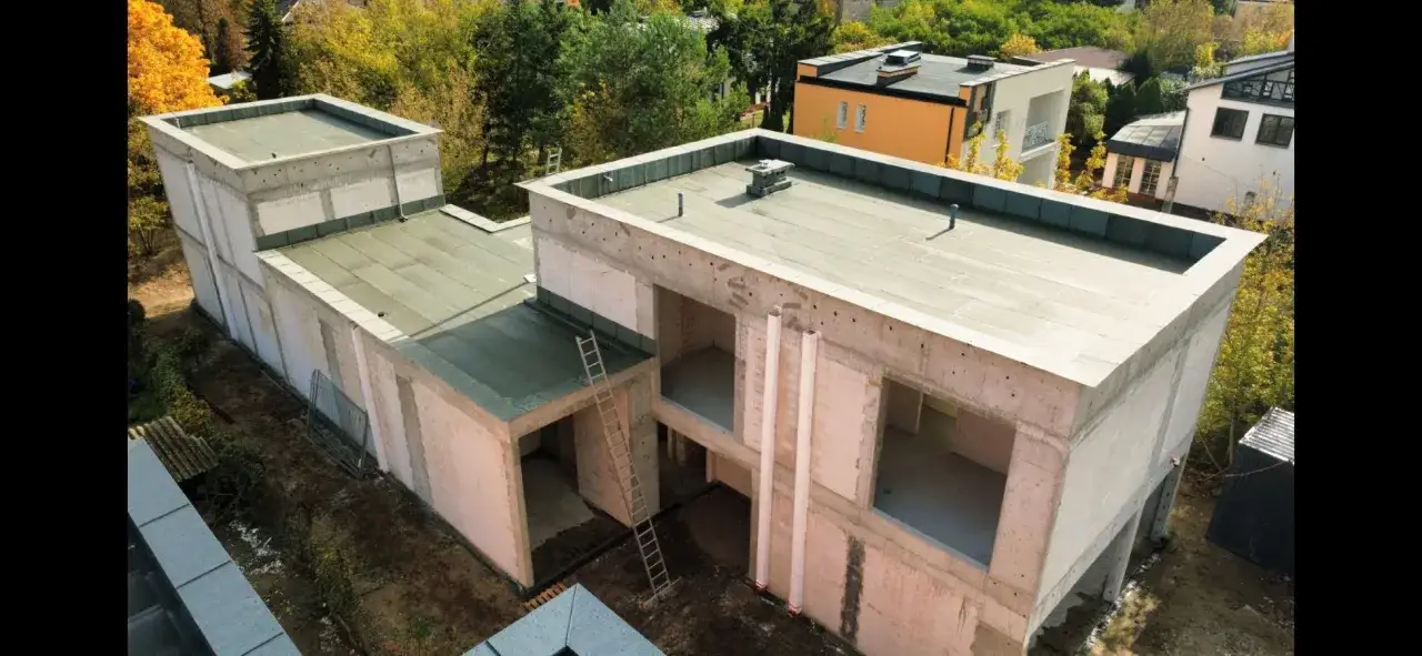 Nowy dom w stanie surowym, z betonowymi ścianami i płaskim dachem, otoczony jesienną zielenią.