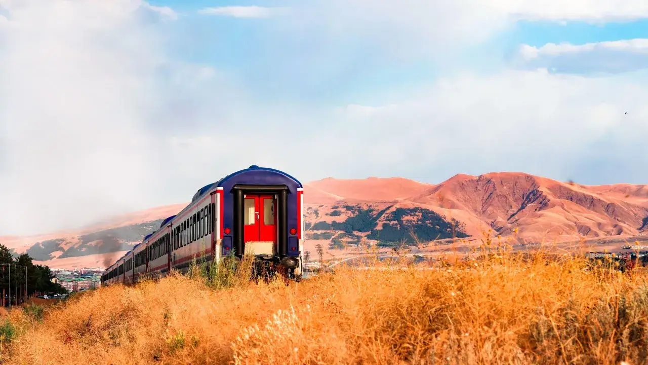 The Bosphorus Express train travels through a dry, golden landscape with rolling, reddish-brown mountains under a cloudy blue sky.