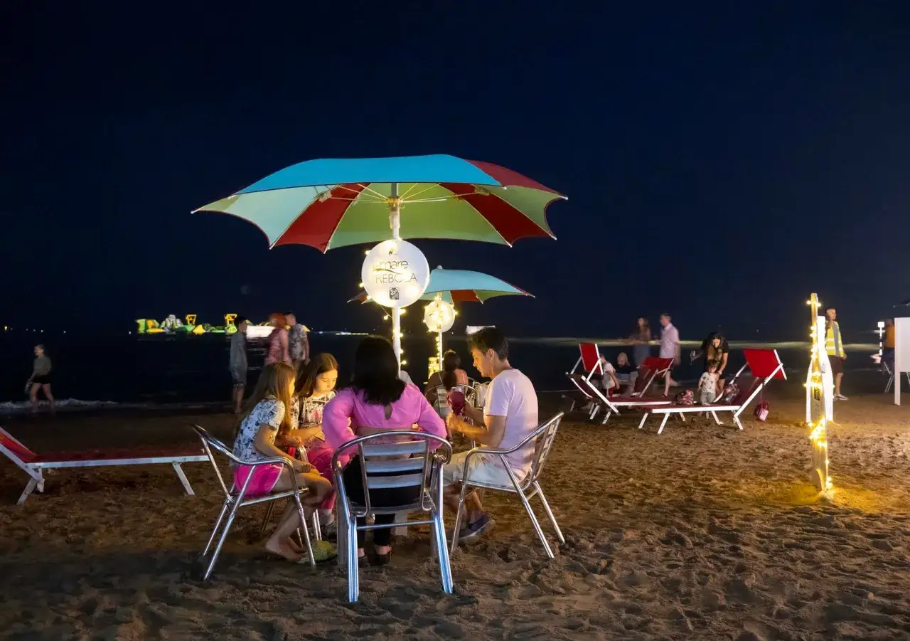 Serata magica alla darsena Rimini: famiglie e amici cenano sotto ombrelloni colorati, illuminati da lucine, mentre il mare fa da sfondo.