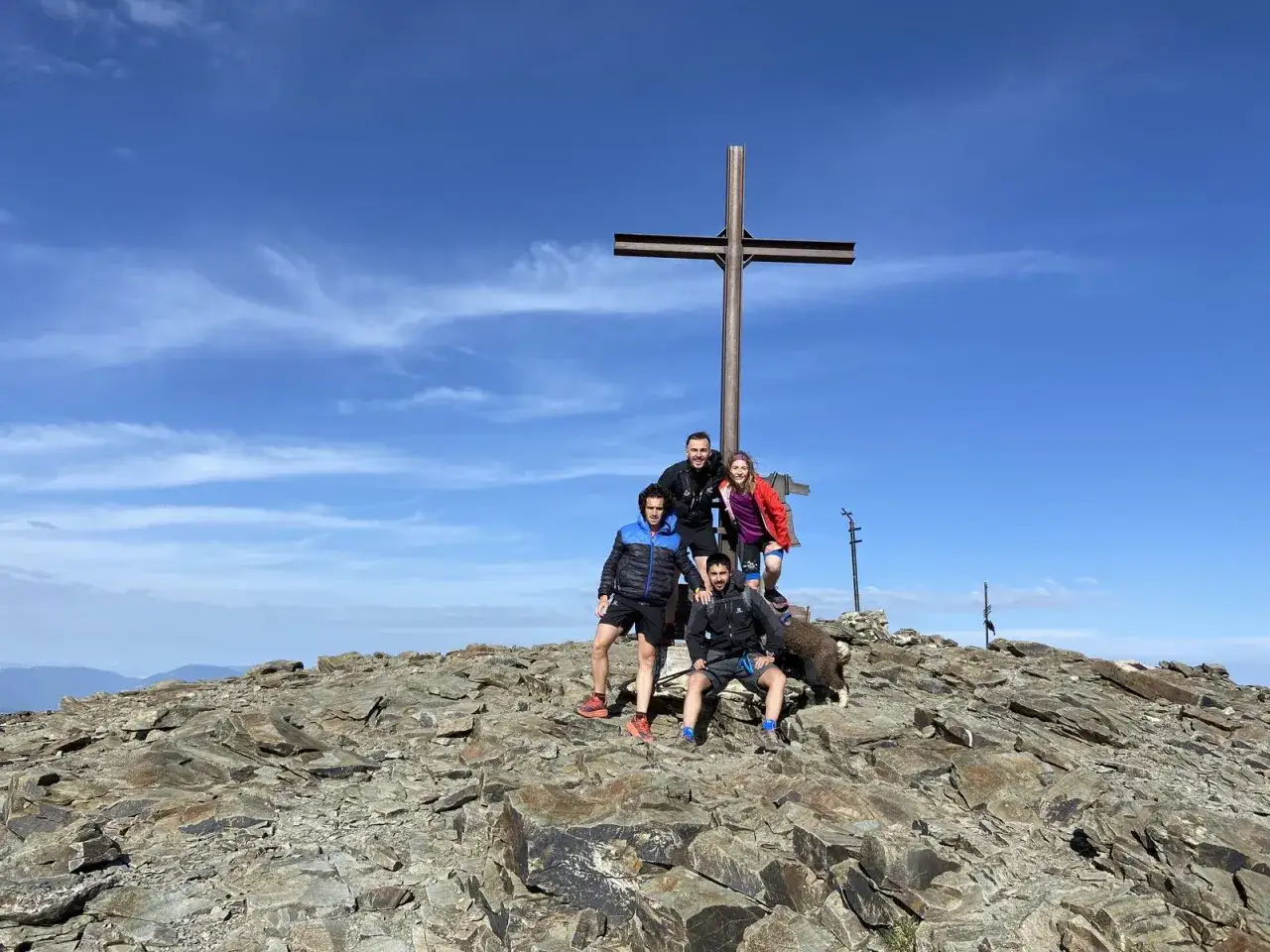 Grupo de amigos en la cima de Fontalba Puigmal, con una cruz met&aacute;lica y un cielo azul despejado.