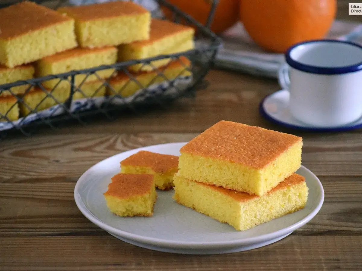 Porción de coca de naranja esponjosa apilada en un plato. En el fondo, más trozos en una cesta y una taza.