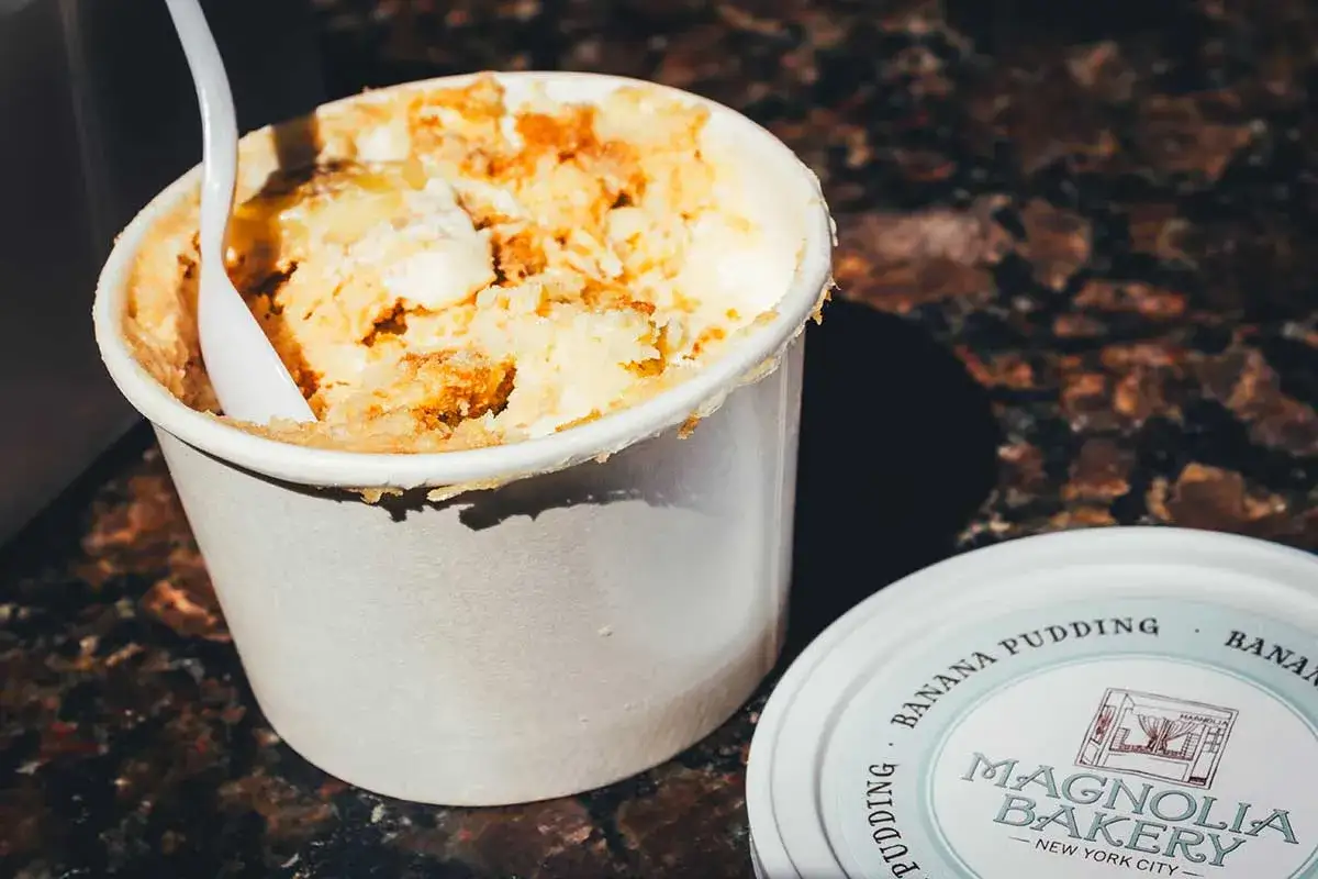 Cremiger Bananenpudding mit zerbr&ouml;selten Keksen, serviert in einem Becher. Ein L&ouml;ffel steckt darin. Das Deckelchen der Magnolia Bakery liegt daneben. Ein tolles Bananenpudding Rezept!