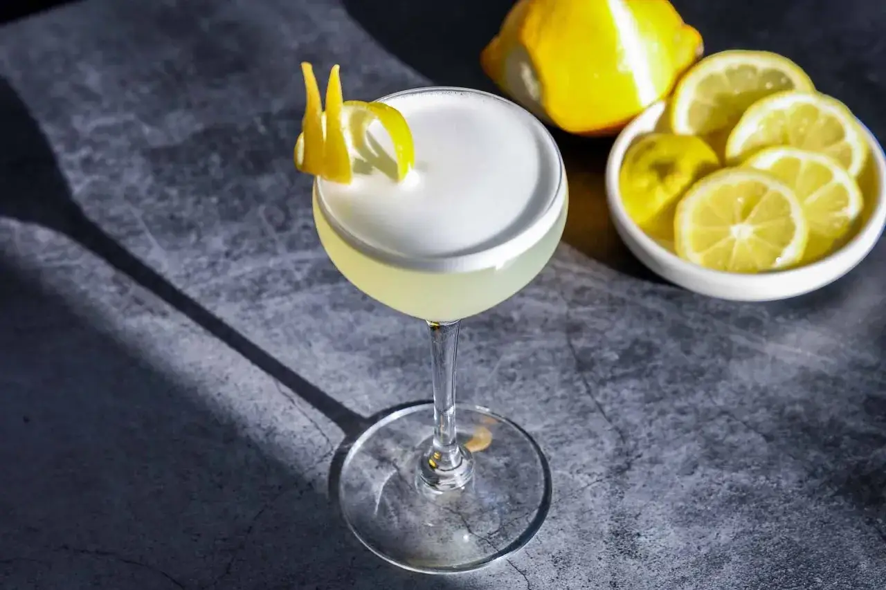 Elegancki drink white lady z pianką i cytrynową ozdobą, obok plasterki cytryny i cała cytryna.