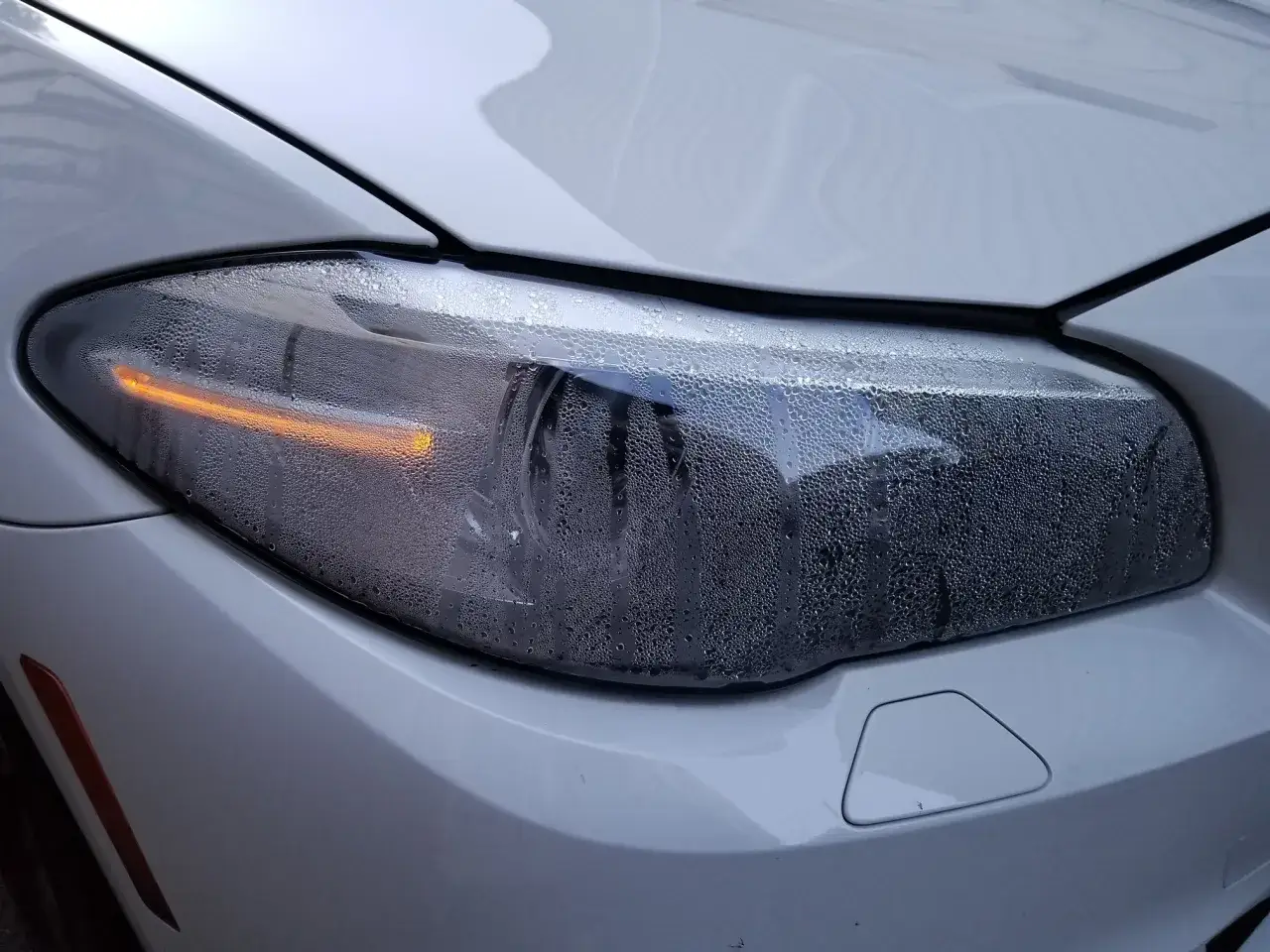 Zaparowane reflektory białego samochodu, z widoczną pomarańczową żar&oacute;wką kierunkowskazu. Krople pary wodnej pokrywają szkło.