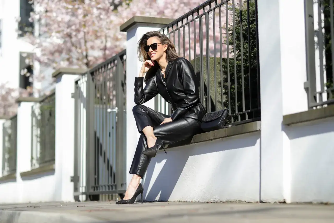 Elegancka kobieta w czarnych sukienkach z ekoskóry, siedząca na murku, z kwitnącą wiśnią w tle.
