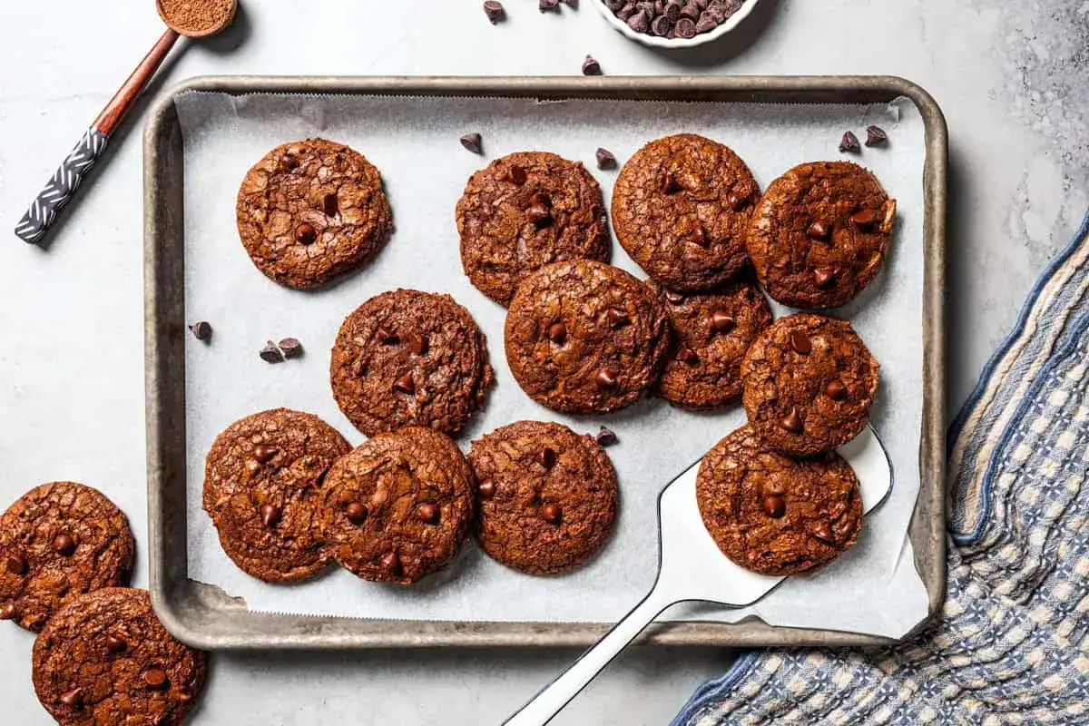 En pl&aring;t med nybakade brownie cookies, rikligt med chokladbitar. En sked med kakaopulver och en sk&aring;l med chokladknappar syns i bakgrunden.