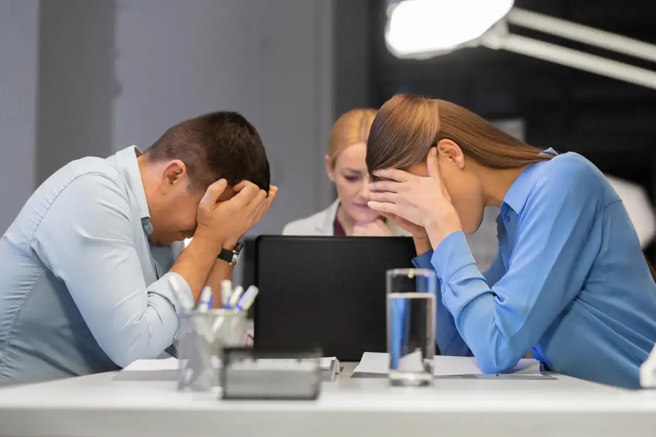 Trois employés accablés, mains sur le visage, face à un écran d'ordinateur. L'ambiance suggère un **mauvais manager** et une surcharge de travail.
