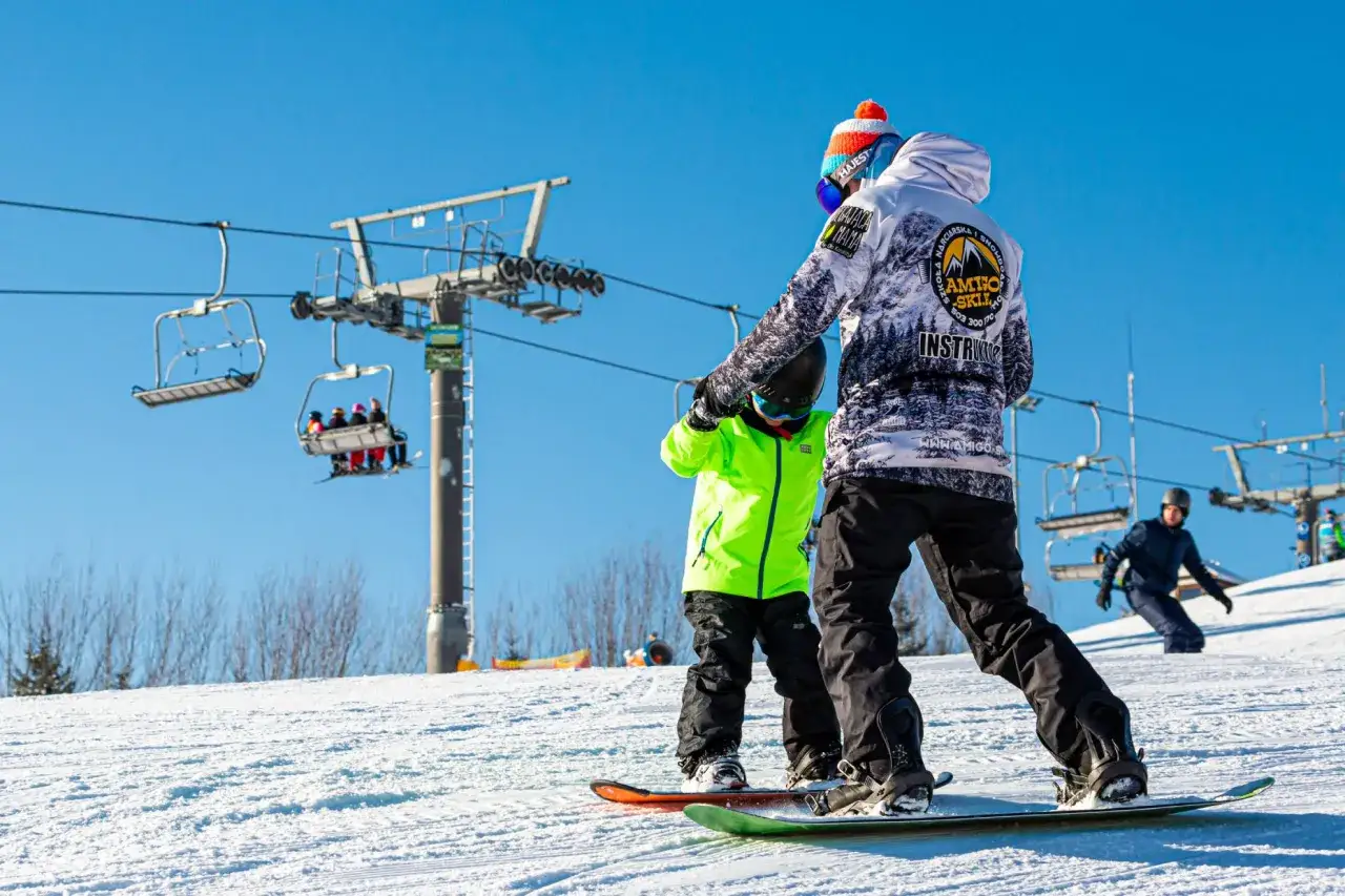 Instruktor pokazuje dziecku, jak jeździć na snowboardzie. Na stoku widać wyciąg krzesełkowy.