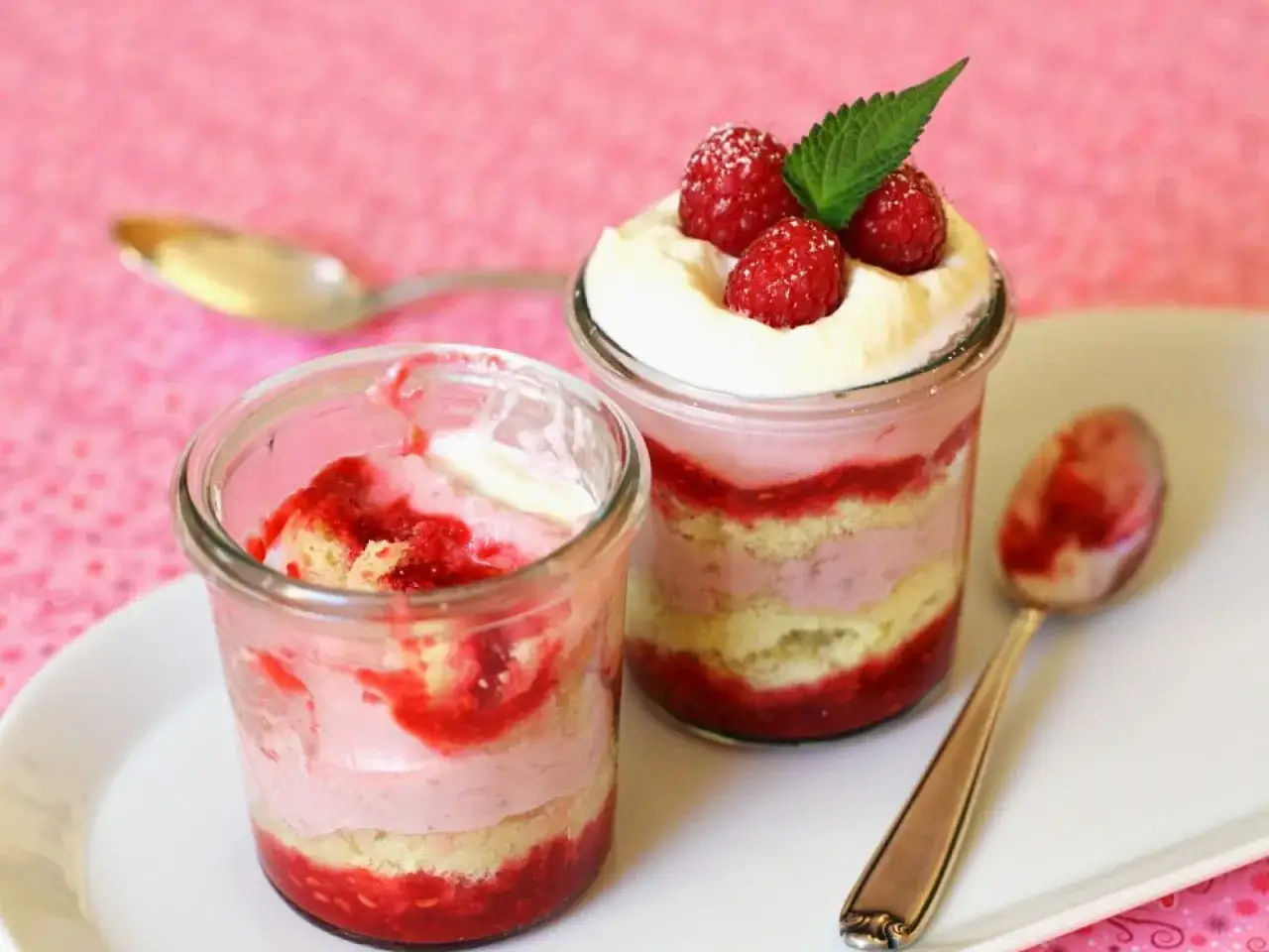 Zwei Gl&auml;ser mit Himbeer-Biskuit-Dessert, geschichtet mit fruchtiger Creme und Sahne. Ein Glas ist bereits angebrochen, das andere ist mit frischen Himbeeren und Minze garniert. Herrlich leichte Sahne Creme!
