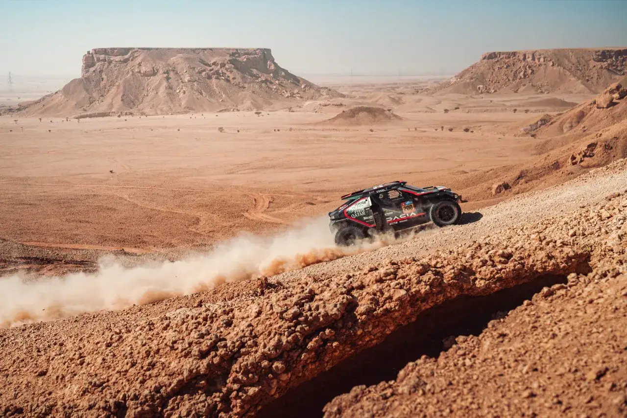 Un coche Dacia Dakar levanta polvo en un paisaje des&eacute;rtico con formaciones rocosas.