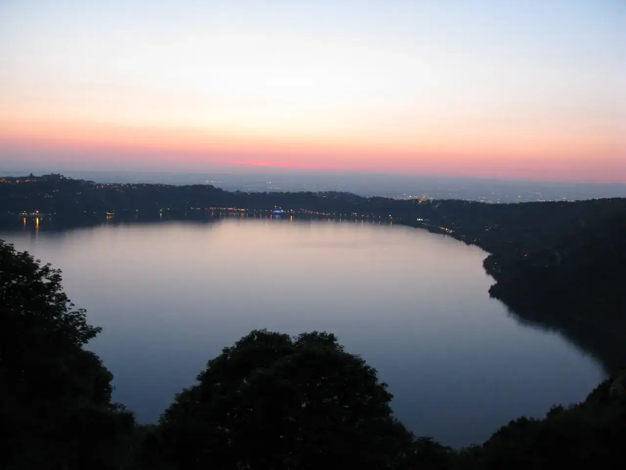 Tramonto sui colli di Roma oggi, con un lago calmo che riflette le luci della città e il cielo colorato.