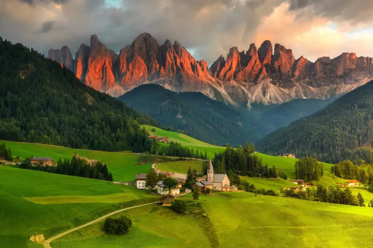 Villaggio alpino nel Val di Funes, con montagne rocciose illuminate dal sole al tramonto e verdi pascoli.