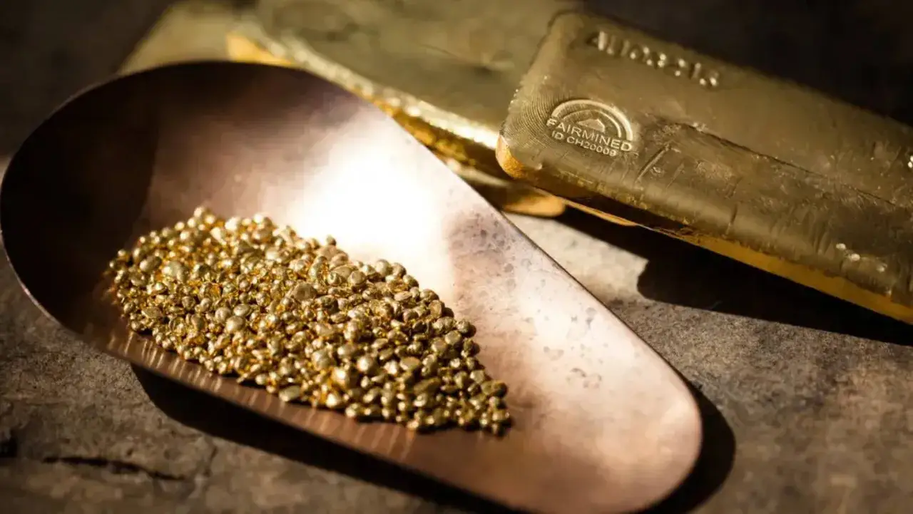 Granelli di oro riciclato in una paletta di rame, con lingotti d'oro sullo sfondo.