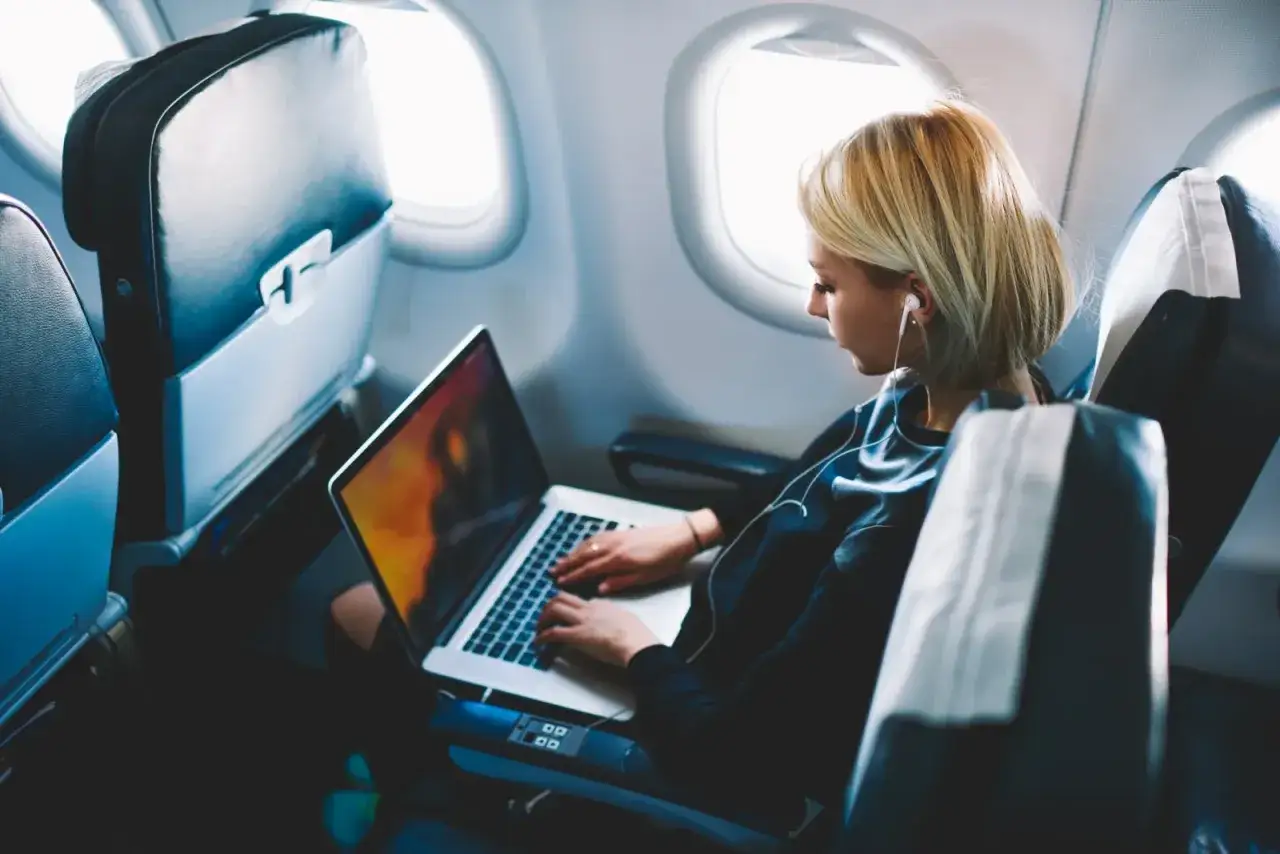 Kobieta pracuje na laptopie w samolocie, słuchając muzyki. To dow&oacute;d, że w samolocie można używać internetu.