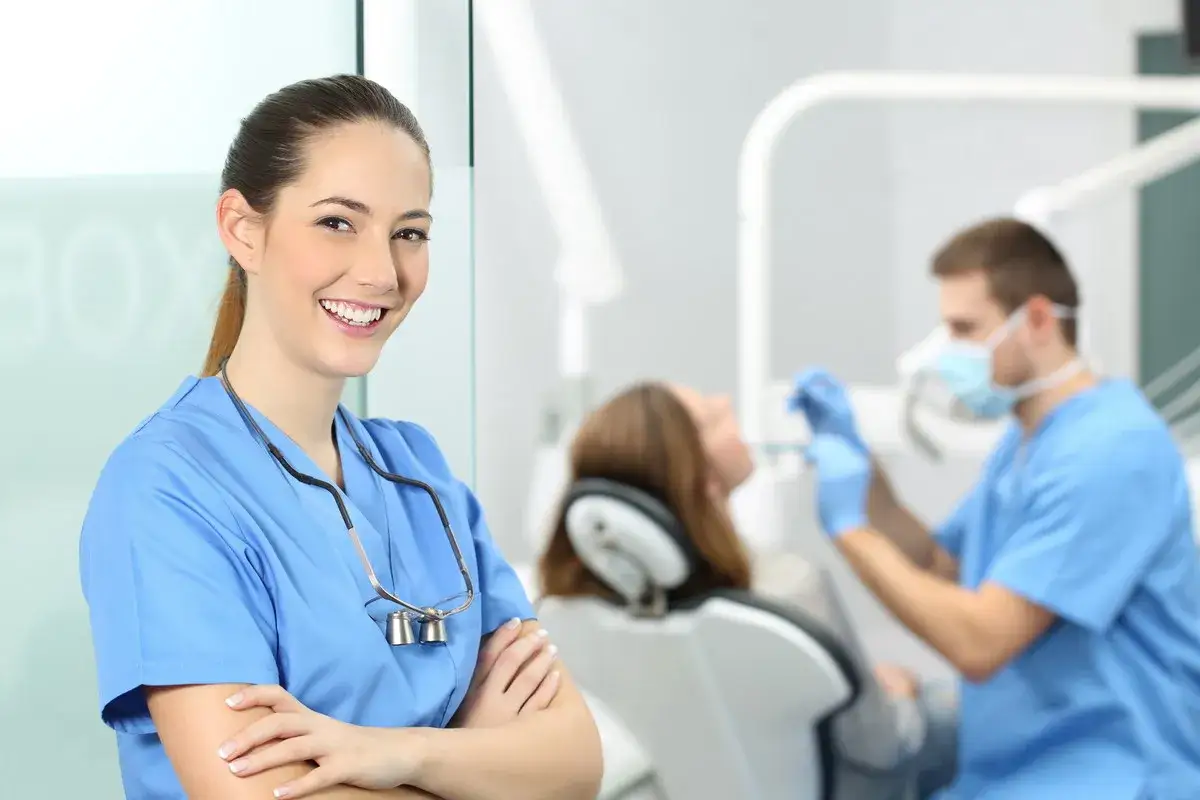 Uśmiechnięta asystentka stomatologiczna w niebieskim uniformie. W tle dentysta z pacjentem. Zastanawiasz się, ile zarabia asystentka stomatologiczna?