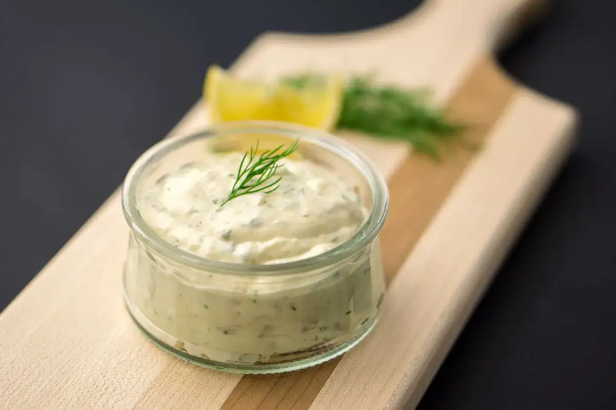 Salsa tartara cremosa in un vasetto di vetro, guarnita con aneto, su un tagliere di legno, con spicchi di limone sullo sfondo.