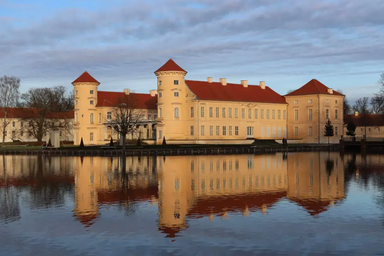 Das Schloss Rheinsberg spiegelt sich im Wasser. Die historische Altstadt von Rheinsberg zeigt sich in gelben Mauern und roten Dächern.