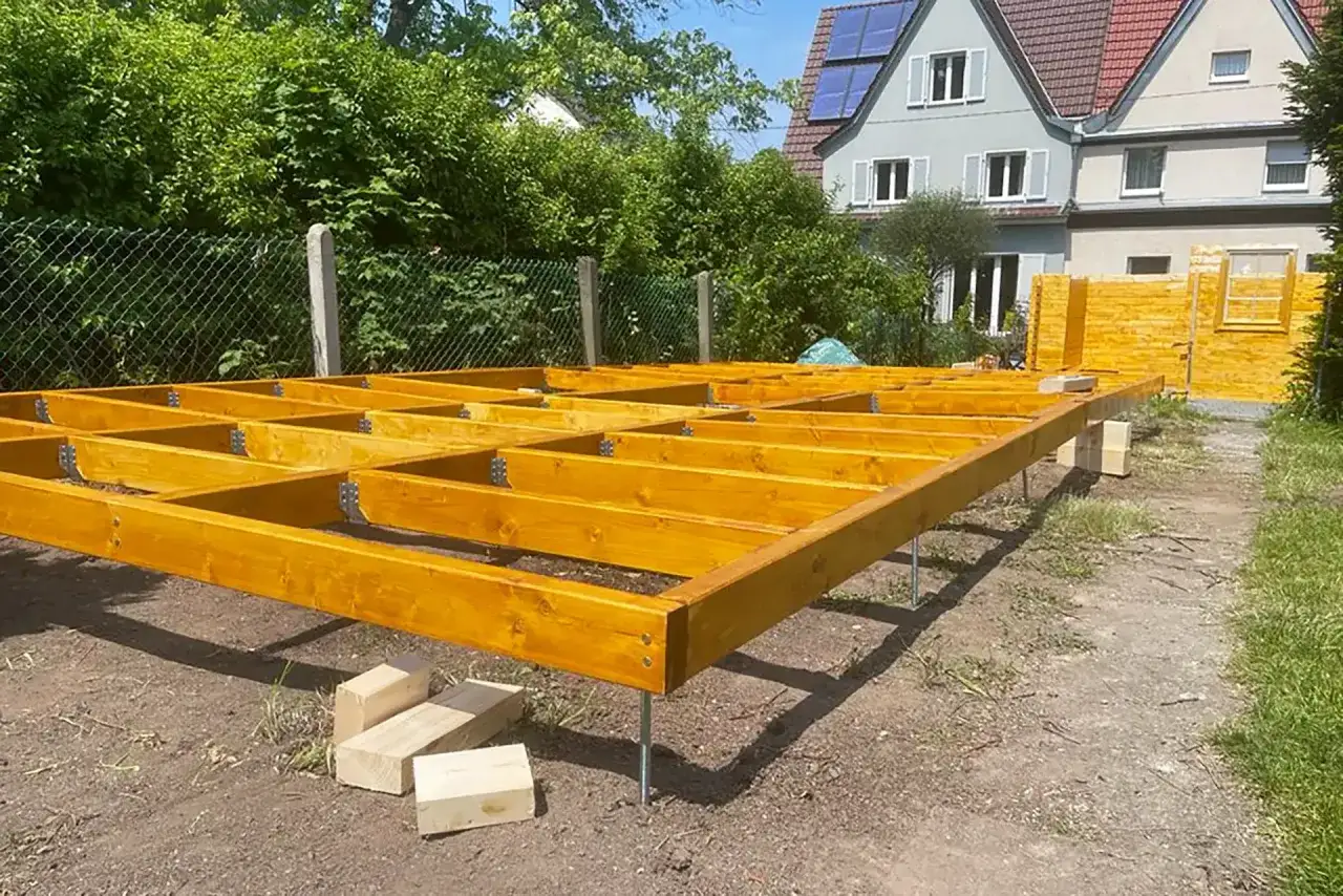 Holzbalken f&uuml;r den Gartenhaus Boden werden auf Pfosten montiert. Ein Teil des Gartenhauses steht bereits im Hintergrund.