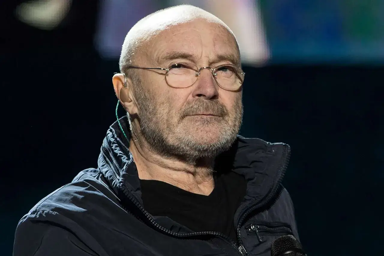 Phil Collins, mimo choroby, w okularach i ciemnej kurtce, z mikrofonem.