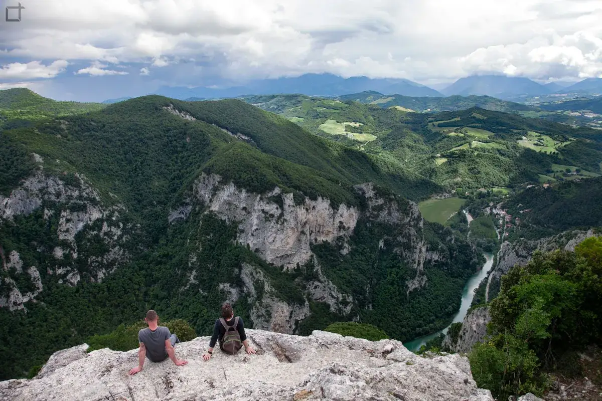 Due escursionisti ammirano la **Gola del Furlo**, un canyon verdeggiante con un fiume turchese che serpeggia tra montagne imponenti.