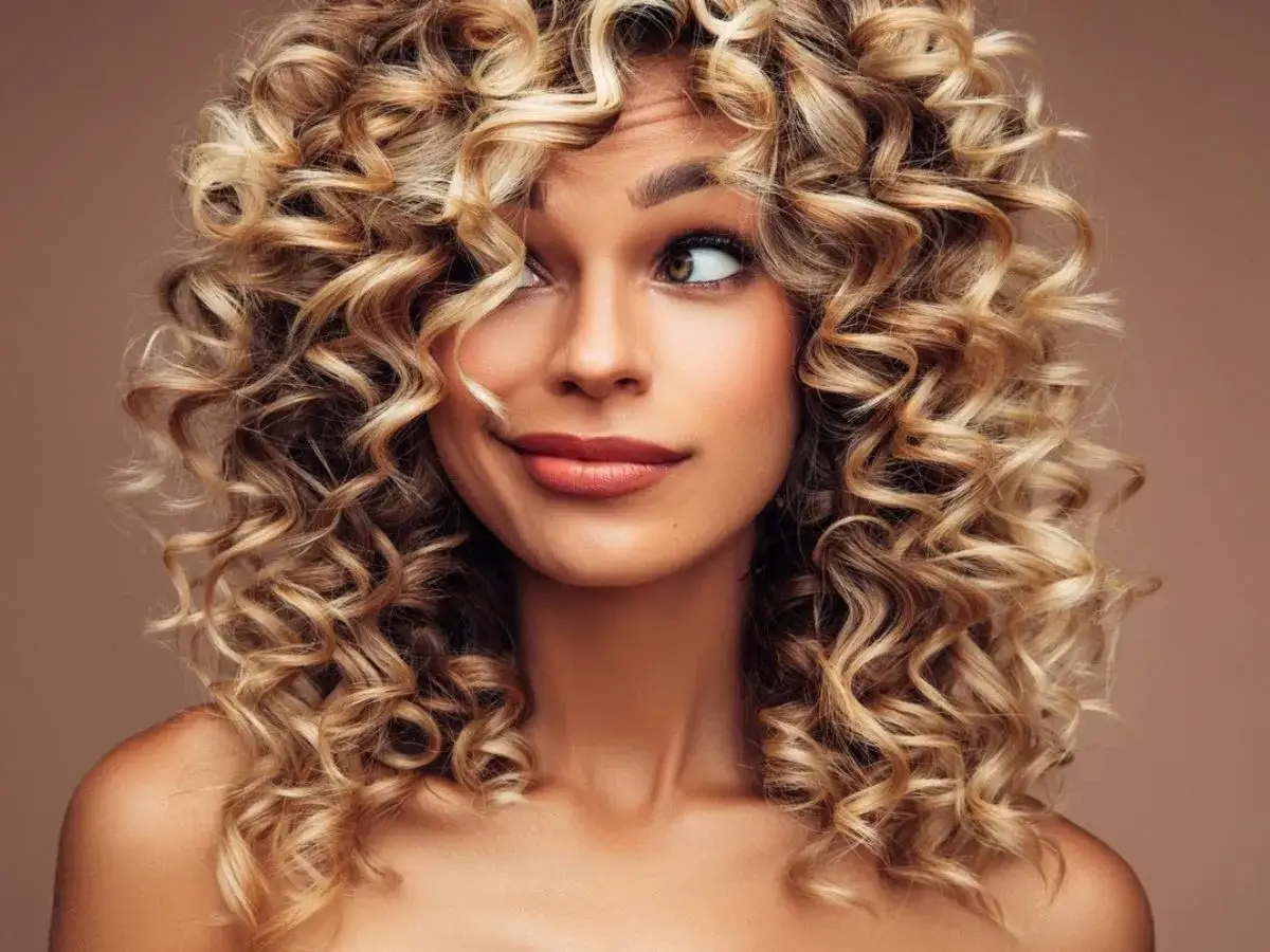 Kobieta z burzą blond, kręcone włosy, patrzy w bok z figlarnym uśmiechem.