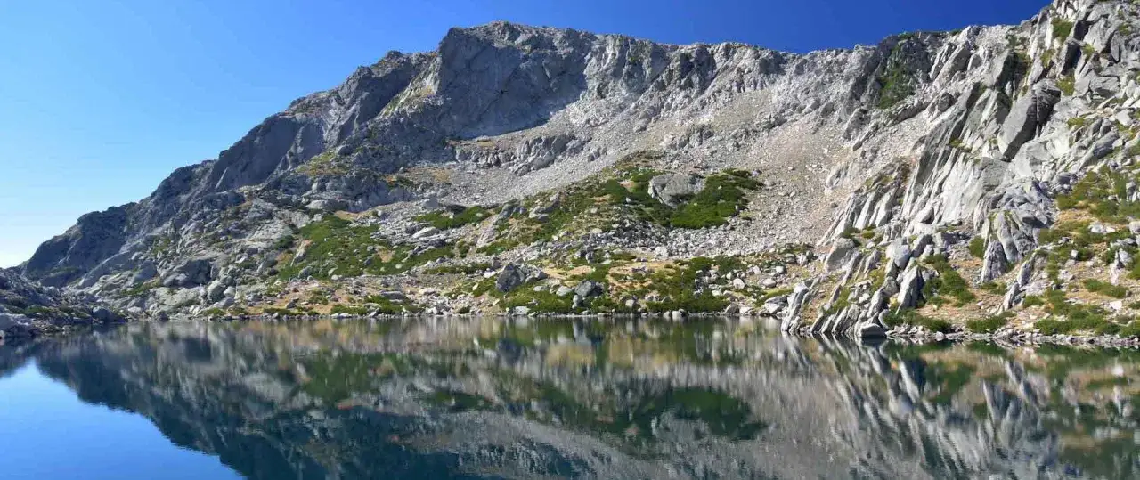 Le lac de Bastani, aux reflets cristallins, est niché au pied d'une montagne rocheuse et escarpée, parsemée de végétation verte.