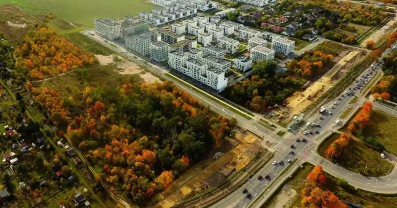 Jesienne barwy lasu otaczają nowe osiedle w Żernikach Wrocławskich. Widać nowoczesne bloki i drogę z ruchem samochodowym.