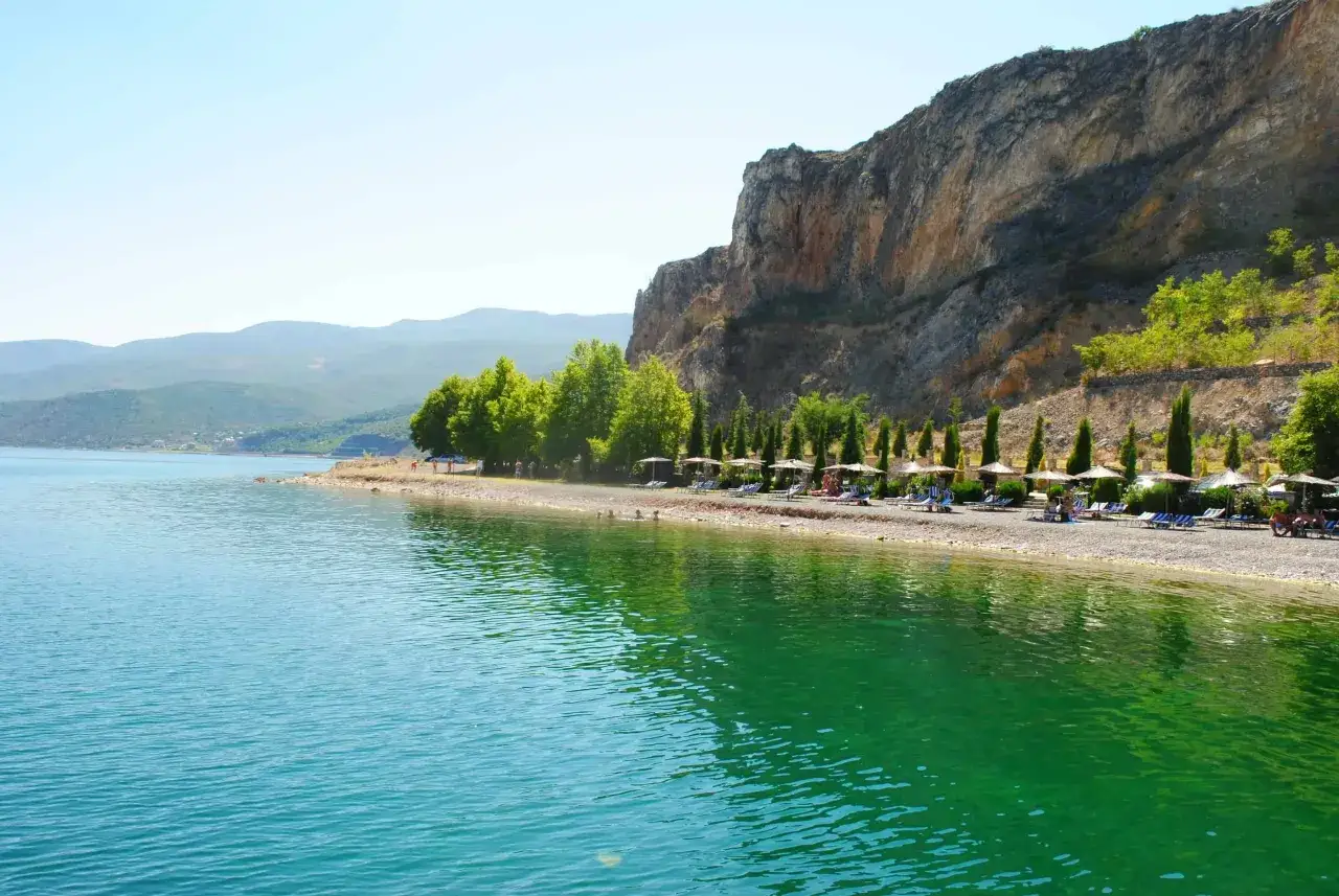 Słoneczna plaża w Pogradec, Albania, z turkusową wodą i skalistym zboczem w tle.