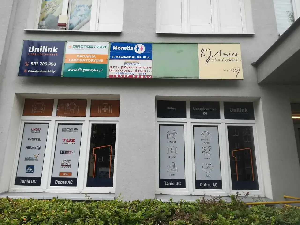 Szyldy firm ubezpieczeniowych i salonu fryzjerskiego na szarej ścianie budynku.