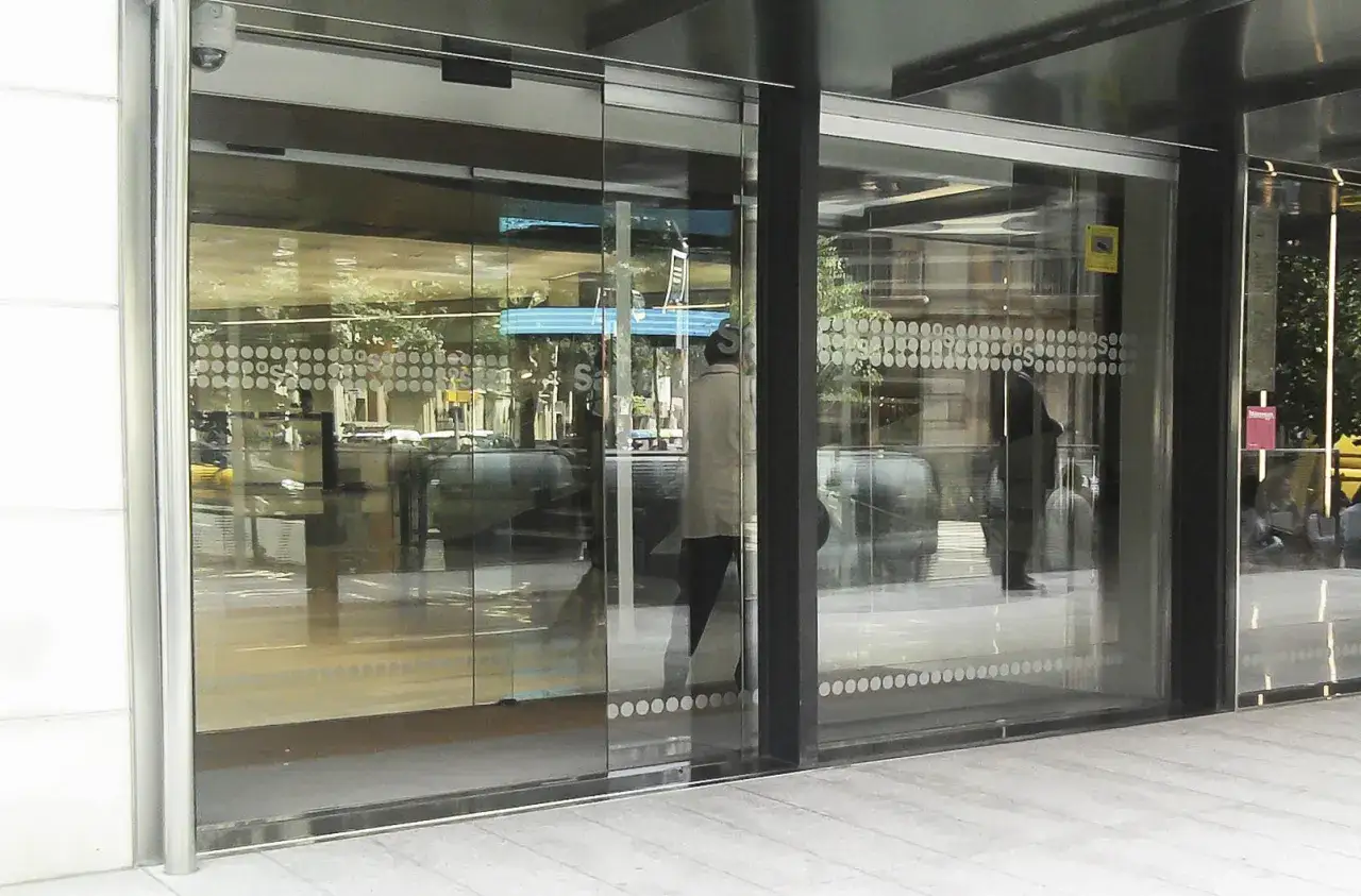 Entrada a un edificio con puertas automáticas de cristal blindado. Reflejos de árboles y edificios en el exterior.