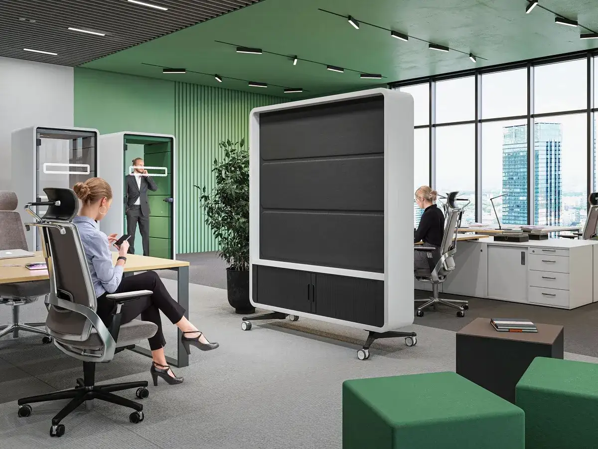 Dans un bureau moderne, une femme regarde son téléphone, assise à son bureau. Un homme en costume parle au téléphone dans une cabine insonorisée. Une scène qui pourrait rappeler le défi de travailler avec son ex.