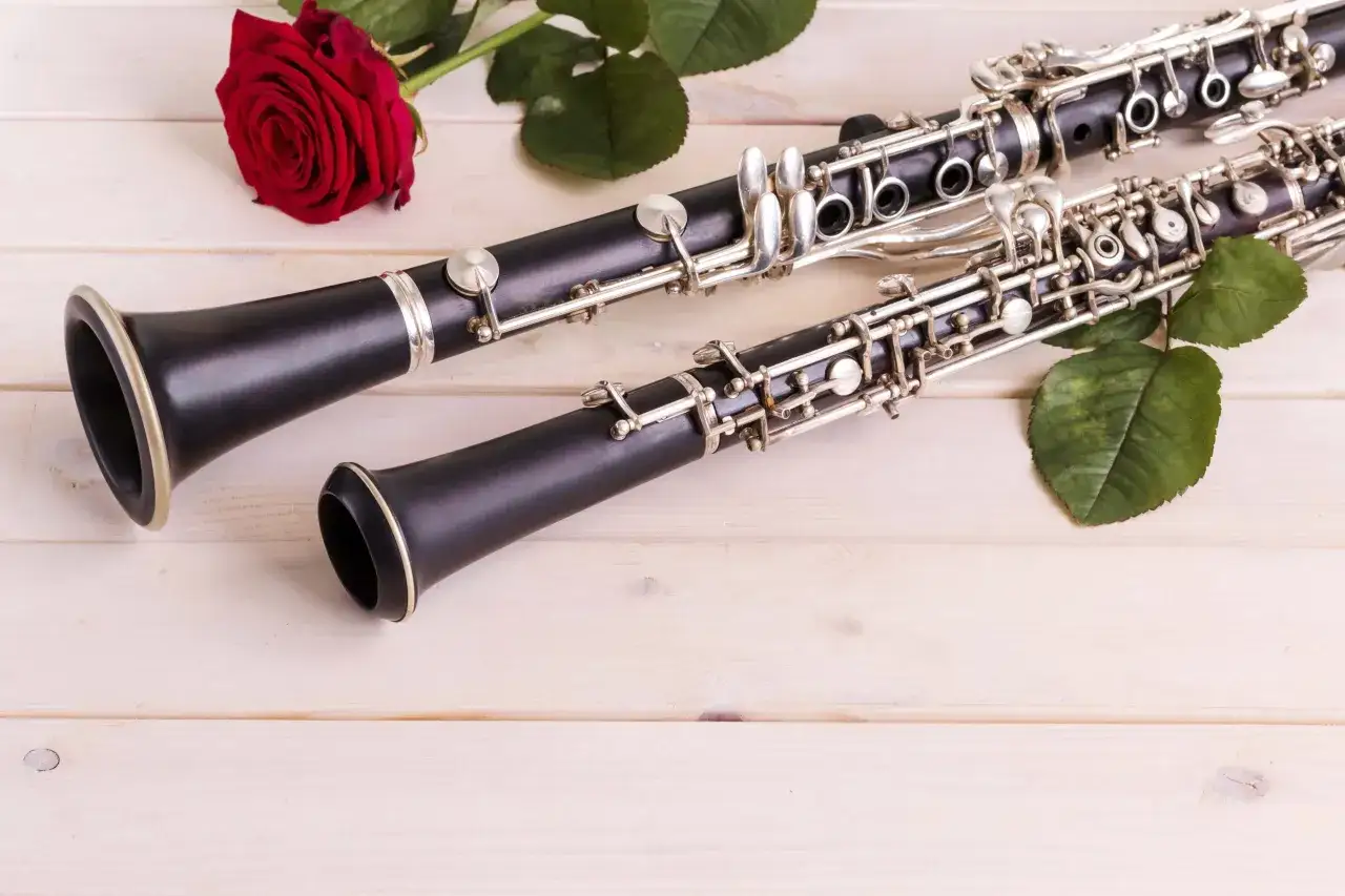 Dwa instrumenty dęte drewniane: obój i klarnet, leżą obok czerwonej róży na jasnym, drewnianym tle.