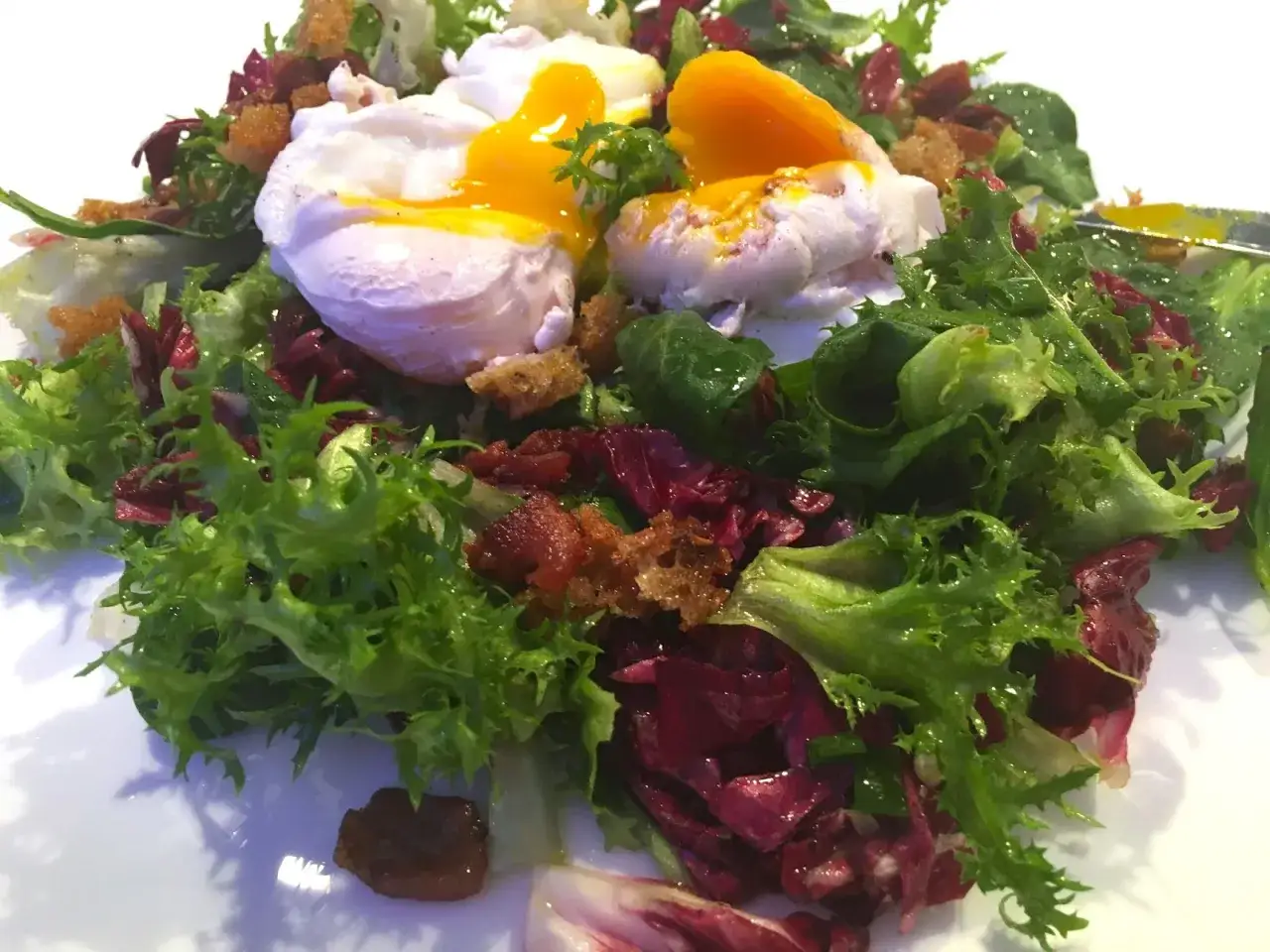 Ein frischer Salat mit knackigem Grün, roten Blättern, knusprigen Croutons und zwei perfekt pochierten **Eiern mit Speck**. Das flüssige Eigelb fließt über die Zutaten.