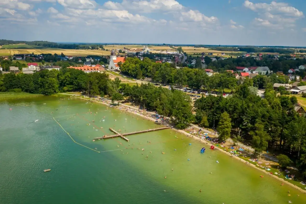 Letnie popołudnie nad jeziorem wielewskim. Tłumy plażowiczów, kąpielisko z pomostem i urokliwa miejscowość w tle.