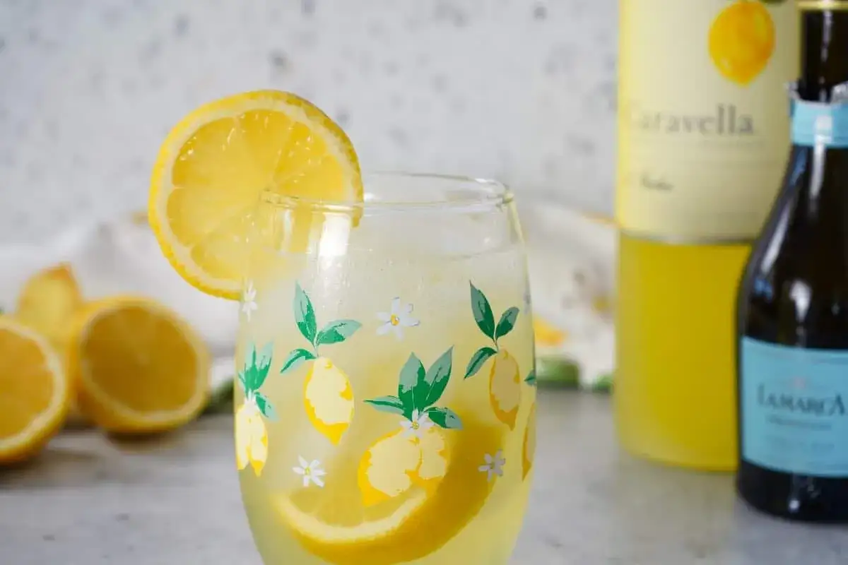 Ett glas med en uppfriskande limoncello drink, garnerad med en citronskiva. Bredvid står flaskor med Caravella och La Marca Prosecco.