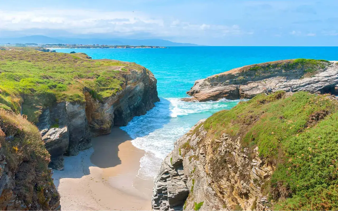 Acantilados verdes y rocosos en la costa artabra, con una playa de arena y el mar azul turquesa.