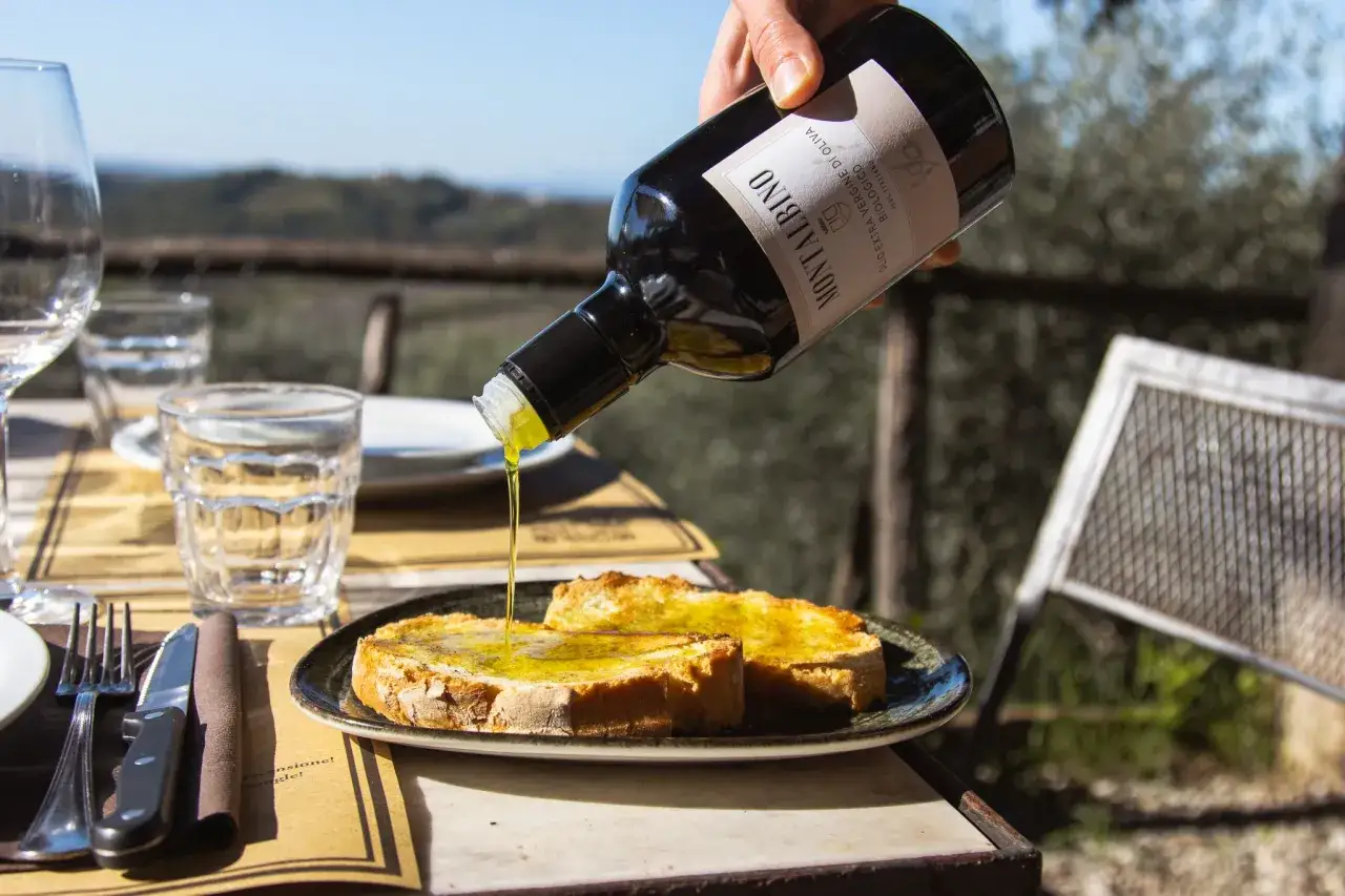 Un filo dorato di **olio di oliva** biologico Montalbino bagna delle fette di pane tostato, su un tavolo all'aperto con vista collinare.