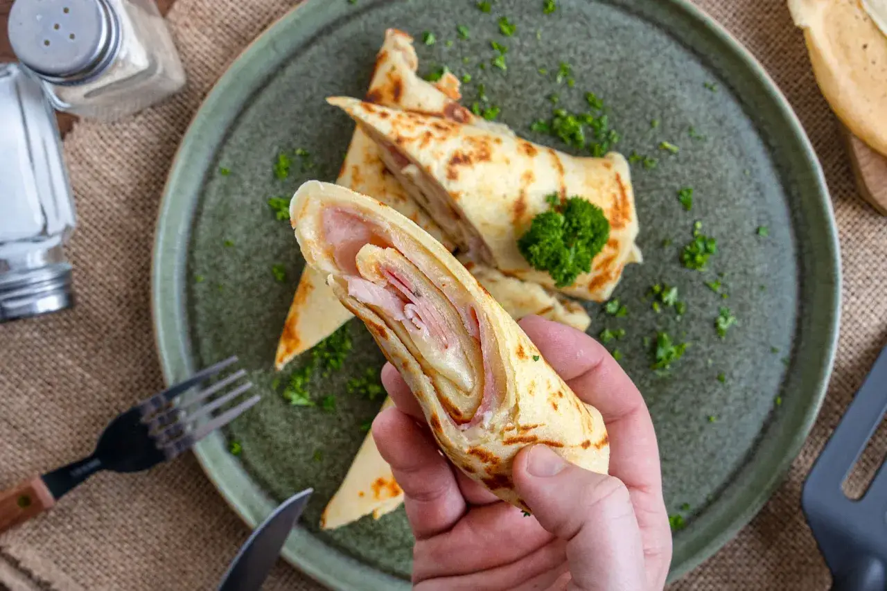 Eine Hand hält einen herzhaften Pfannkuchen, gefüllt mit Schinken und Käse. Auf dem Teller liegen weitere gerollte Pfannkuchen, garniert mit Petersilie.
