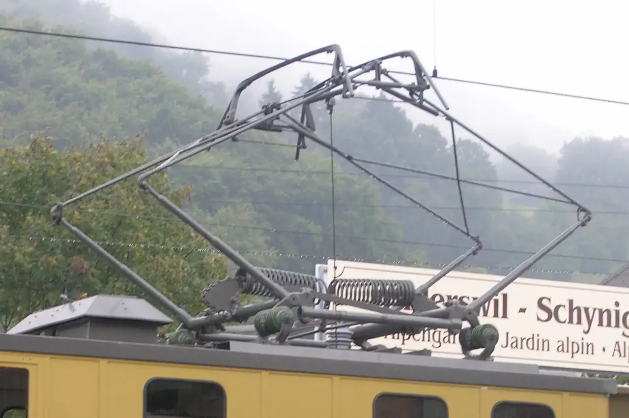 Złożony mechanizm sieci trakcyjnej na dachu żółtego pociągu, gotowy do pobierania prądu z przewodów.